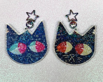 May include: A pair of iridescent blue cat earrings with red and white eyes and a star charm. The earrings are made of clear acrylic and have a glittery finish.