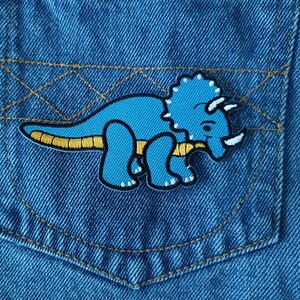 May include: Close-up of a blue denim pocket with a cartoon triceratops patch. The dinosaur is light blue with white horns and a yellow belly. The pocket has gold stitching.
