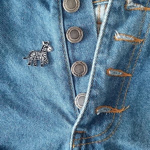 May include: Close-up of a light blue denim garment with silver buttons and a small zebra pin. The buttons are embossed with a circular design. The denim has visible stitching and a worn appearance.