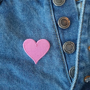 May include: A close-up of a pair of blue denim jeans with a pink heart-shaped patch sewn on the front. The jeans have silver metal buttons.