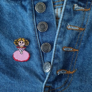 May include: Close-up of blue denim jeans with silver button closures. A small, embroidered patch of a cartoon princess in a pink dress is on the jeans. The jeans have visible stitching and a classic button-fly design.