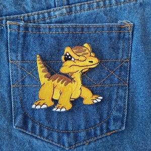 May include: A yellow embroidered dinosaur patch with brown stripes and black eyes, sewn onto a blue denim pocket.