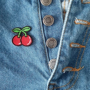 May include: Close-up of blue denim fabric with silver buttons and a cherry patch. The patch features two red cherries with green leaves. The buttons are round with a textured design.
