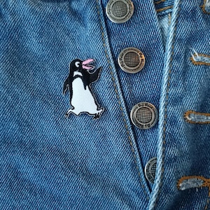 May include: A black and white penguin patch with a pink beak sewn onto a blue denim jacket. The patch is centered on the jacket and is surrounded by silver buttons.