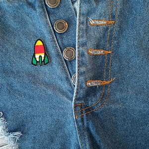 May include: A close-up of a pair of blue denim jeans with a rocket ship patch on the front. The jeans have a button fly and are distressed with a small rip on the left leg.