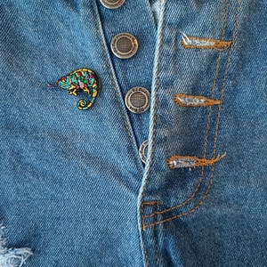 Peut inclure: Gros plan sur un short en jean bleu avec une épingle caméléon colorée. Le short a des fermetures à boutons argentés et des détails usés. L'épingle caméléon présente une variété de couleurs, dont le vert, le jaune et le bleu.