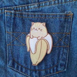 May include: A blue denim pocket featuring an embroidered patch of a cat inside a banana. The patch uses beige, tan, white, and brown thread. The pocket has yellow stitching.