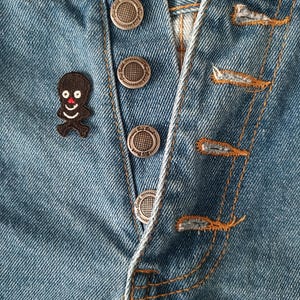 May include: Close-up of blue denim jeans with a button fly. The jeans feature silver metal buttons and orange stitching. A small black patch with a smiling face is sewn onto the jeans.