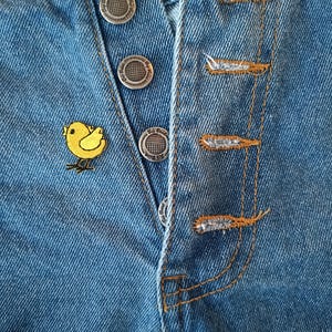 May include: Close-up of blue denim shorts with a yellow chick patch. The shorts have silver buttons and orange stitching. The denim is faded in areas, giving it a worn look.