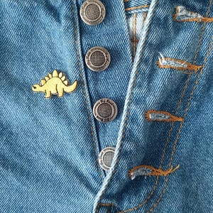 May include: Close-up of light blue denim jeans with silver buttons and a yellow dinosaur pin. The jeans have visible stitching in a contrasting orange-brown thread. The buttons are embossed with a circular design.