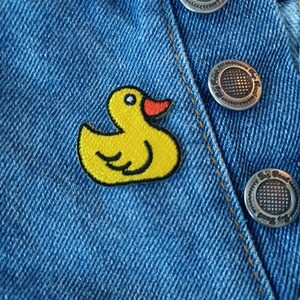 May include: A yellow rubber ducky patch sewn onto a blue denim jacket. The duck has a red beak and black eyes.