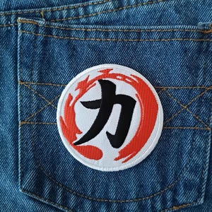 May include: A round patch with a white border, featuring a black Japanese character within a red and orange flame-like design. The patch is sewn onto the back pocket of a blue denim garment.