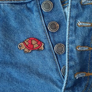 May include: Close-up of a blue denim garment with silver metal buttons. A small, embroidered turtle patch with a brown body and a red shell is sewn onto the denim. The garment's seams are highlighted with yellow stitching.