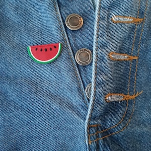 May include: Close-up of a blue denim garment with silver buttons and orange stitching. A watermelon slice patch is sewn on the denim. The garment has a button-up closure.