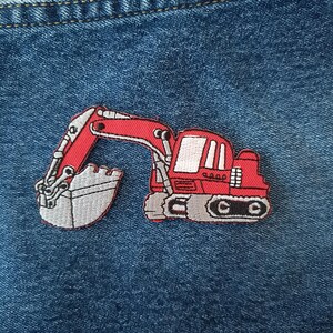 May include: A red embroidered excavator patch with a grey bucket and tracks, set against a blue denim background. The patch features white detailing and is likely for clothing or accessories.