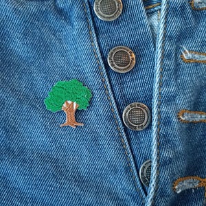 May include: A close-up of a pair of blue denim jeans with a green embroidered tree patch and silver metal buttons.
