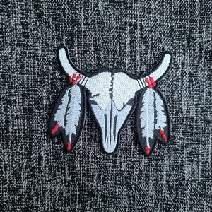 May include: White embroidered patch of a bull skull with two white feathers on each side. The feathers have red tips. The skull has red accents on the horns.