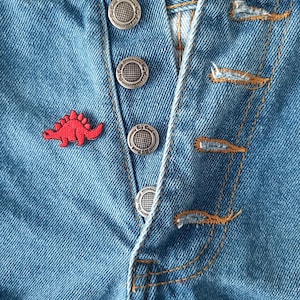 May include: Close-up of blue denim jeans with a button fly. The jeans have four silver-colored buttons and a small red dinosaur patch on the front. The stitching is in a contrasting orange color.
