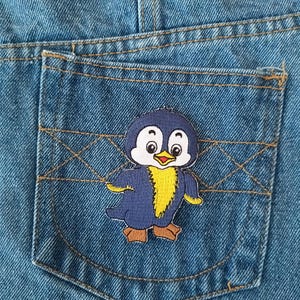May include: Close-up of a pair of blue denim jeans with a small pocket featuring a cartoon penguin patch. The penguin has a blue body, yellow chest, and brown feet. The jeans have visible stitching.