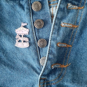 May include: Close-up of a light blue denim garment with silver buttons and orange stitching. A pink and white carousel pin is attached to the denim. The buttons are circular with a textured design.