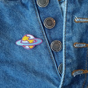 May include: A blue denim jacket with silver buttons and a colorful embroidered patch of a planet with a ring around it.
