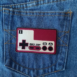 May include: Close-up of a blue denim pocket with a rectangular patch featuring a retro video game controller design. The patch has a burgundy background with white and black details, including the text "SELECT START" and buttons.