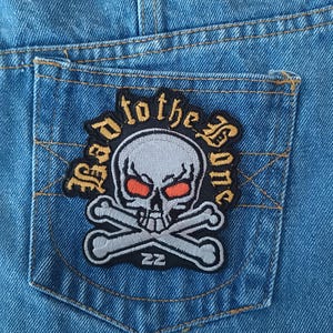 May include: Close-up of a blue denim pocket with a patch. The patch features a skull with orange eyes and crossed bones, surrounded by the words "Dead to the Bone" in a gothic font. The patch has a black border.