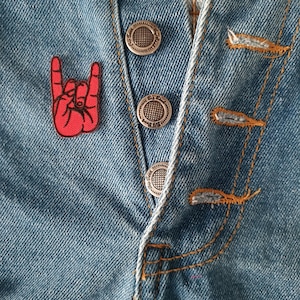 May include: Close-up of a light blue denim garment with silver metal buttons and orange stitching. A red embroidered hand gesture patch is on the left side. The garment has a button-up closure.