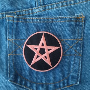 May include: Close-up of a light blue denim pocket with a circular patch. The patch features a pink pentagram on a black background, surrounded by a pink border. The pocket has yellow stitching.
