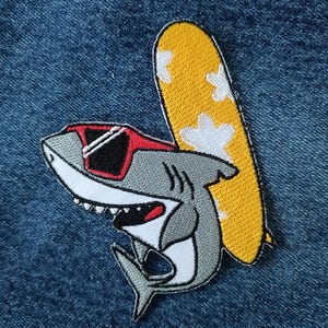 May include: Embroidered patch of a cartoon shark wearing sunglasses and carrying a yellow surfboard with white cloud details. The shark is grey, white, and red, with a smiling expression. The patch is on a blue denim background.