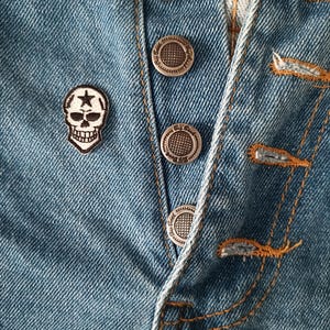May include: Close-up of a light blue denim garment with silver metal buttons and orange stitching. A small skull patch with a star is sewn on the denim. The buttons are embossed with a circular pattern.