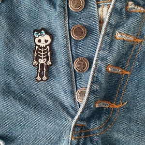 May include: Close-up of blue denim jeans with a decorative patch of a skeleton wearing a blue bow. The jeans have silver metal buttons and orange stitching.