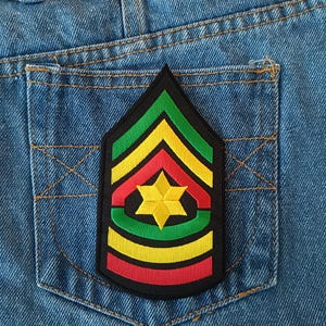 May include: A black, green, yellow, and red embroidered patch in the shape of a military rank insignia, featuring a yellow star in the center. The patch is sewn onto the back pocket of a pair of blue denim jeans.