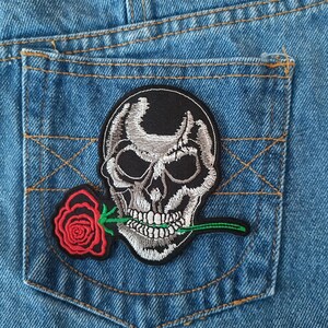 May include: Close-up of a blue denim pocket with a black patch. The patch features a detailed, embroidered skull with a red rose in its mouth. The skull is gray and white, with green stitching on the rose stem.