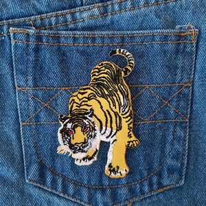May include: An embroidered tiger patch in yellow and black on a blue denim pocket.