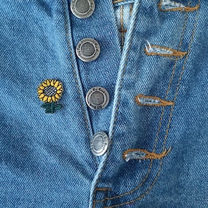 May include: Close-up of a blue denim jacket with silver buttons and an embroidered sunflower.