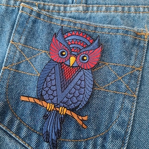 Könnte beinhalten: Nahaufnahme einer blauen Jeans-Tasche mit einem gestickten Eulen-Aufnäher. Die Eule ist blau mit roten und gelben Akzenten und sitzt auf einem braunen Ast. Die Tasche ist mit gelbem Faden genäht.