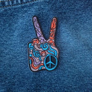 May include: Embroidered peace sign patch with a blue, orange, and purple design. The patch features a peace symbol in the center and is set against a denim background. The patch is a symbol of peace and love.
