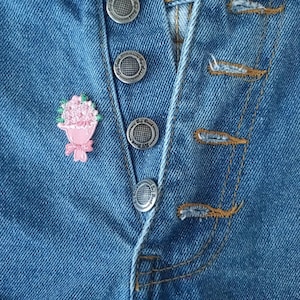 May include: Close-up of blue denim jeans featuring silver metal buttons and a small pink floral patch. The jeans have a button fly and visible stitching.