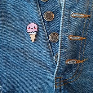 May include: Close-up of a blue denim garment with silver buttons and a small ice cream cone patch. The patch features a pink ice cream scoop with a smiling face atop a brown cone. The garment has visible stitching and distressed details.