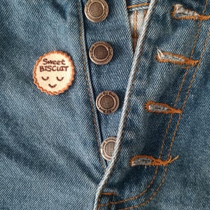 May include: Close-up of blue denim jeans with silver buttons and orange stitching. A small, round patch with the text "Sweet Biscuit" and a smiling face is sewn onto the jeans. The jeans are unbuttoned.