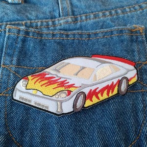 May include: Close-up of a blue denim pocket with a car patch. The patch features a white race car with yellow and red flames on the front and sides. The car has brown wheels and a red spoiler.