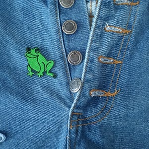 May include: A pair of blue denim jeans with a green frog patch and silver buttons.