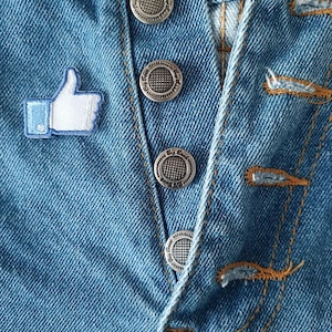 May include: Close-up of blue denim jeans with a thumbs-up patch and silver buttons. The jeans have a button fly and orange stitching. The thumbs-up patch is white with a blue outline.