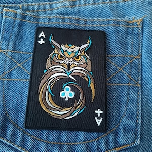 Könnte beinhalten: Ein schwarzer Aufnäher mit einem Eulen-Design in Braun, Gold und Türkis. Der Aufnäher hat die Form einer Spielkarte, mit dem Symbol des Kreuz-Asses und dem Buchstaben "A" sichtbar. Der Aufnäher befindet sich auf einer Jeans-Tasche.