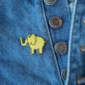 May include: A yellow embroidered elephant patch on a blue denim background with silver metal buttons.