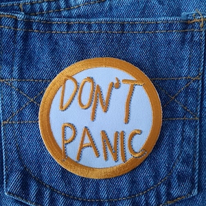Peut inclure: Gros plan sur une poche de jean bleue avec un écusson circulaire. L'écusson présente un fond blanc avec les mots "DON'T PANIC" en orange, soulignés d'or. Le denim présente des coutures visibles.