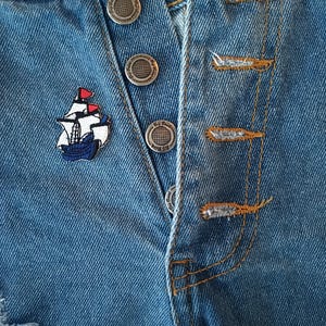 May include: Close-up of blue denim shorts with silver metal buttons and a small embroidered ship patch. The shorts have a distressed look with visible stitching and a button fly.