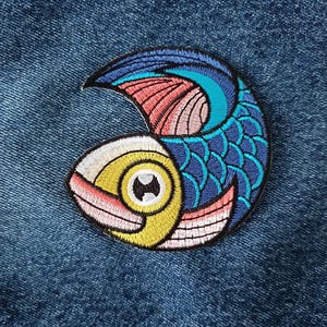 May include: A blue, pink, and yellow embroidered patch of a stylized fish with scales. The fish is in a crescent shape.