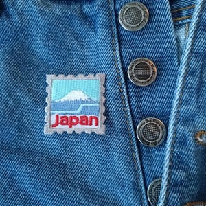 May include: A close-up of a blue denim fabric with a small embroidered patch that says "Japan" and depicts a mountain range. The fabric has several silver buttons.